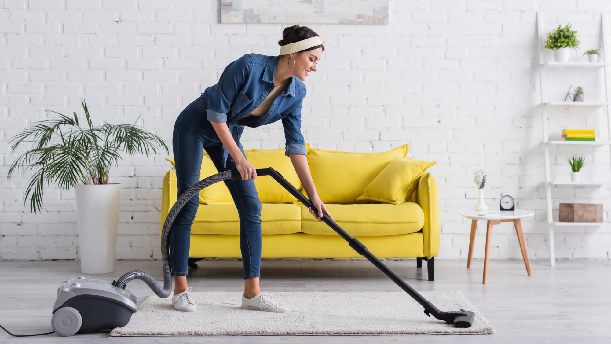 A woman in a blue denim shirt and headband is vacuuming a light-colored carpet in a bright living room. A yellow sofa is positioned against a white brick wall, and there are potted plants and a small table with a clock nearby.