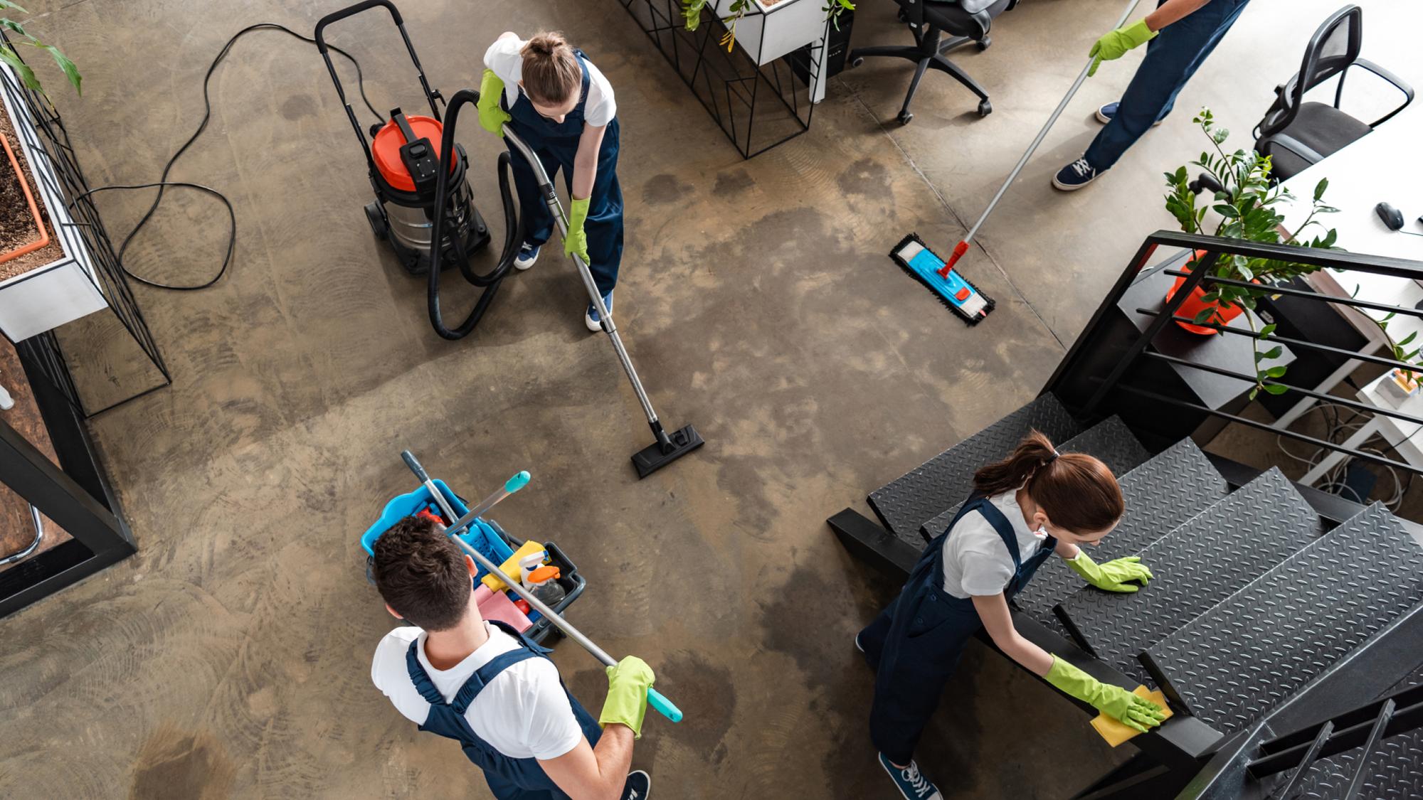 Aerial view of a cleaning crew in a modern office space, with two individuals using mops and a vacuum cleaner, and one person cleaning stairs. All are dressed in blue overalls and wearing yellow gloves. The environment features plants and contemporary furniture.